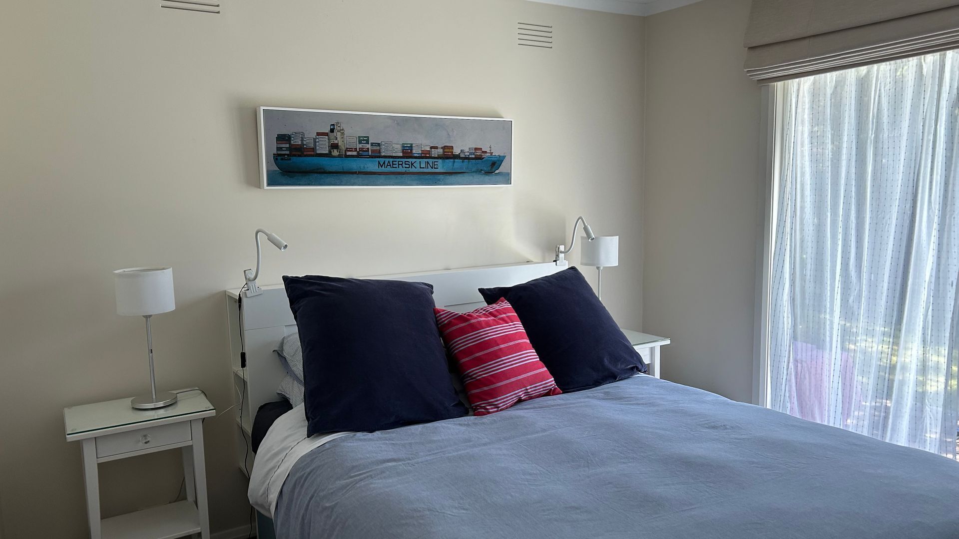 Een net opgemaakt bed met een blauw-witte deken, twee marineblauwe kussens en een rood-witte gestreepte kussen. Boven het bed hangt een lijst met een foto van een vrachtschip. Aan beide zijden van het bed staan nachtkastjes met lampen.