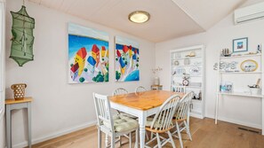 A dining area with a wooden table and white chairs, two colorful paintings on the wall, a green hanging net, and a white shelf with various items.