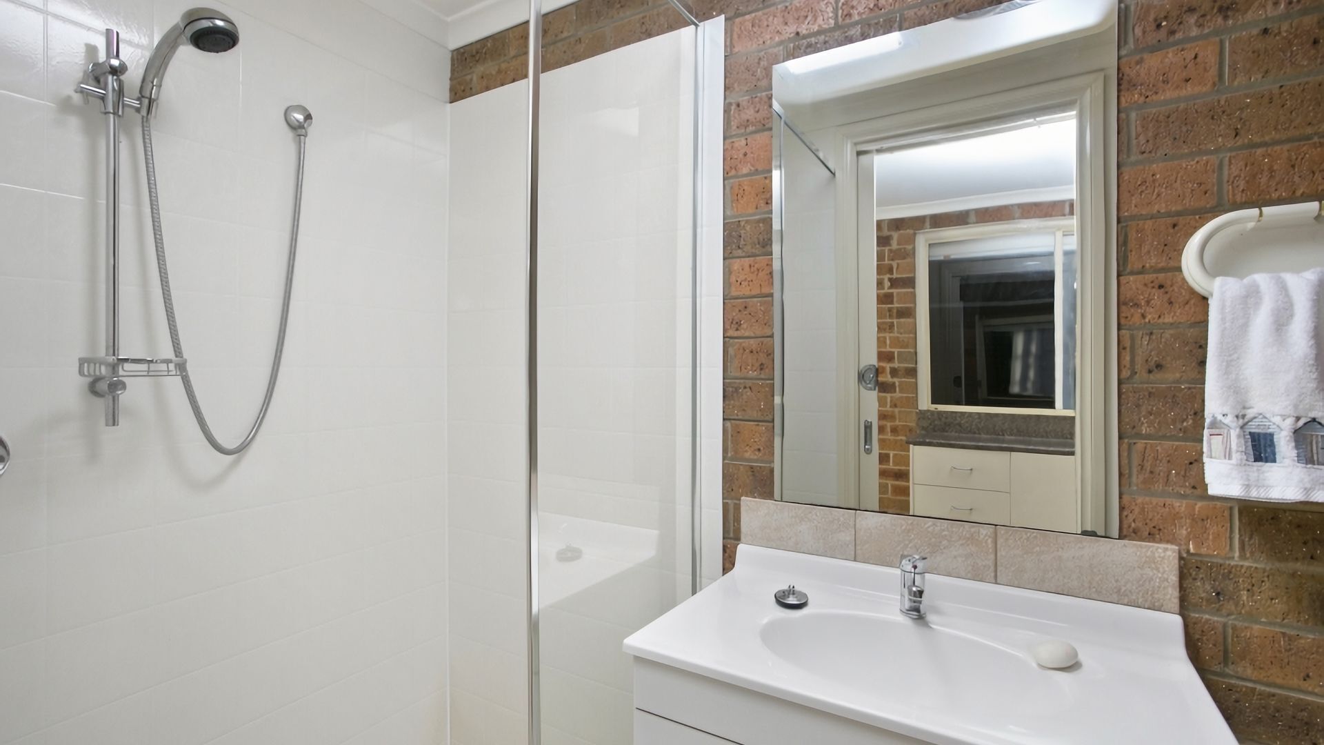 A bathroom with a glass shower enclosure, a white sink, and a mirror reflecting a brick wall.