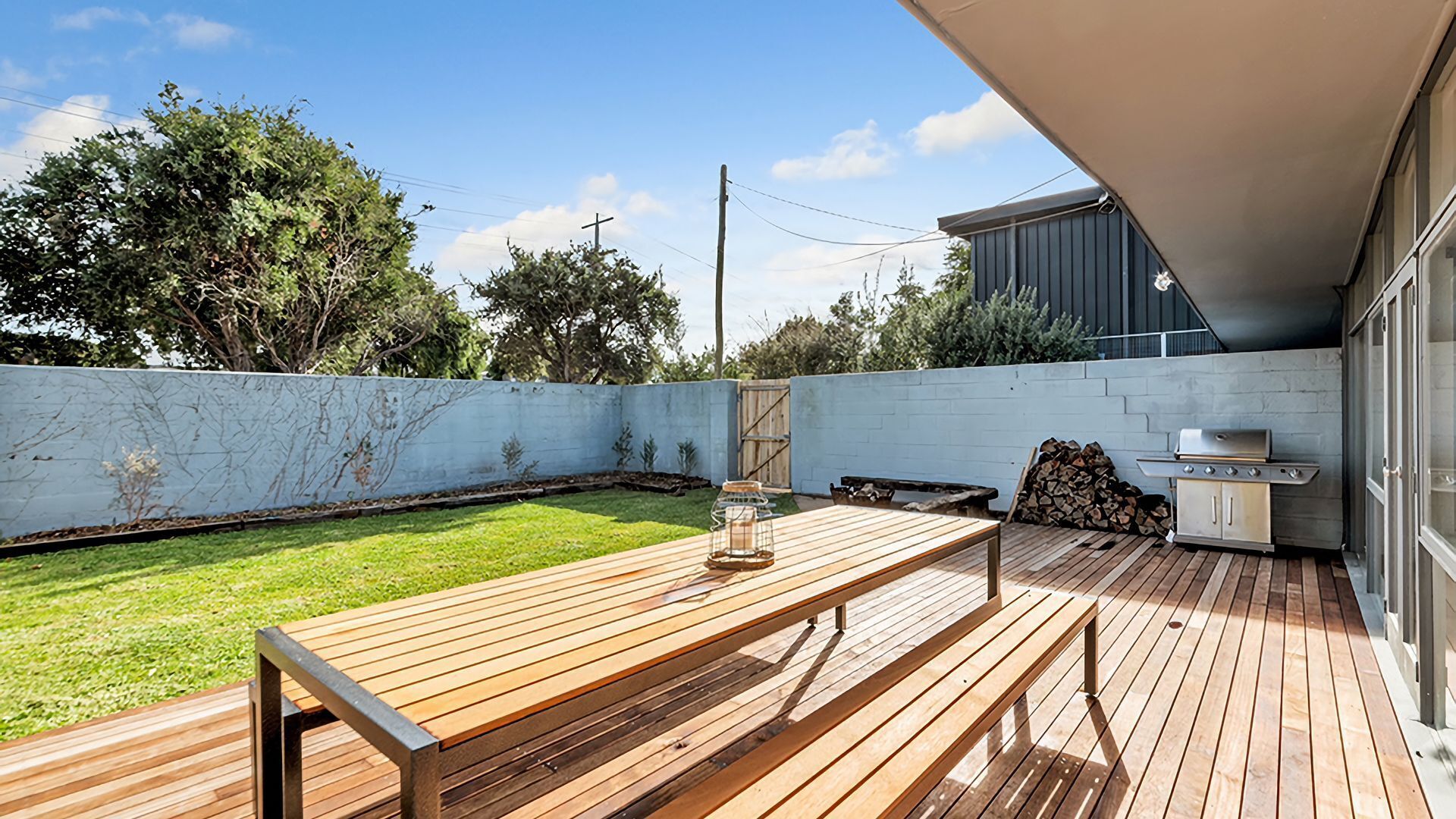 A wooden deck with a grill and a table, a grassy yard, and a white wall with a gate.