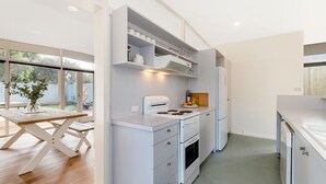 A modern kitchen with white cabinetry, a built-in oven, and a refrigerator. There is a dining table with a vase of flowers and a view of the outdoors through large windows. - The 20th Hole - Point Lonsdale (POINT LONSDALE)