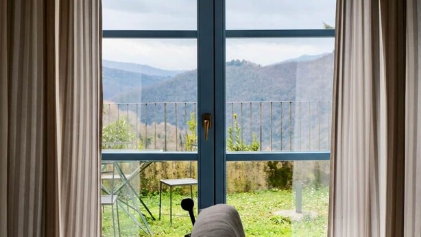 A room with a large window, a view of a mountainous landscape, and a balcony with a chair.
