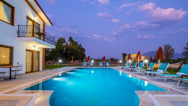 A swimming pool with lounge chairs and umbrellas, surrounded by a building with a balcony and outdoor seating. - Ada Villa (Fethiye)