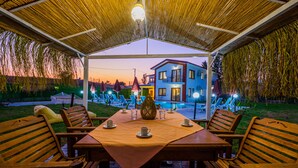 An outdoor dining area with a table set for four, surrounded by wooden chairs, under a thatched roof. - Ada Villa (Fethiye)