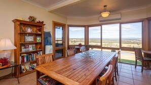Un coin repas avec une table et des chaises en bois, une bibliothèque remplie de livres et d’objets décoratifs, et de grandes fenêtres offrant une vue sur le paysage.