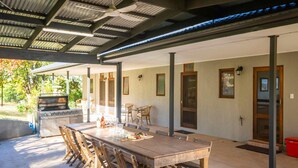 Une terrasse extérieure avec une longue table et des chaises en bois, un barbecue et un espace abrité doté d’un toit en métal.