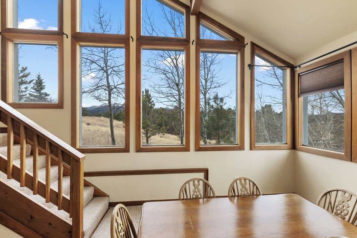 A dining area with a wooden table and chairs, large windows with a view of trees and mountains, and a wooden staircase.