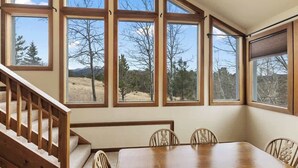 Un comedor con mesa y sillas de madera, ventanas amplias con vista a árboles y montañas, y una escalera de madera.