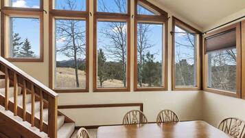 A dining area with a wooden table and chairs, large windows with a view of trees and mountains, and a wooden staircase.