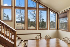 A dining area with a wooden table and chairs, large windows with a view of trees and mountains, and a wooden staircase.