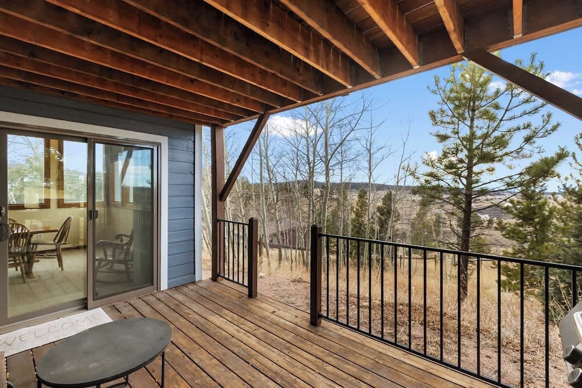 Um deck de madeira com porta de vidro que dá para um quarto, guarda-corpo de metal e vista para árvores e uma montanha.