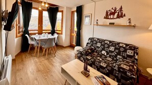 Una sala de estar acogedora con suelo de madera, un sofá estampado, una mesa de comedor con sillas y vistas al exterior a través de amplios ventanales.