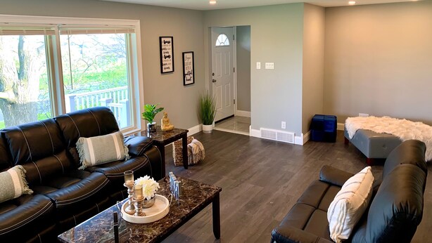 A spacious living room with a dark leather couch, a coffee table with decorative items, and a view of the outdoors through large windows. - The Grand Gorge Villa-With Yard& Parking (Niagara Falls)