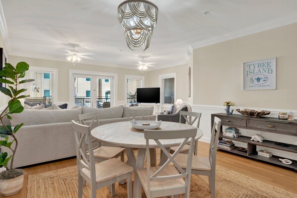 A living room with a round table, white chairs, a grey sofa, a chandelier, and a TV.