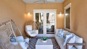 A sunlit patio with a ceiling fan, wicker chair, white sofa, and a small table with a plant. - Great Family Condo with Pool Views! Pool Access (Tybee Island)