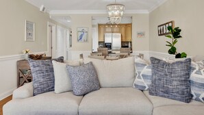 A living room with a sofa, pillows, a chandelier, and a kitchen in the background. - Great Family Condo with Pool Views! Pool Access (Tybee Island)