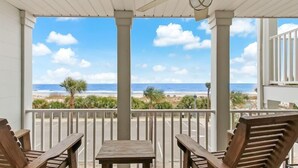 A covered outdoor area with a table and two chairs, overlooking a beach with the ocean and a clear sky.
