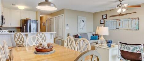 A dining area with a wooden table and white chairs, a kitchen in the background, and a ceiling fan.