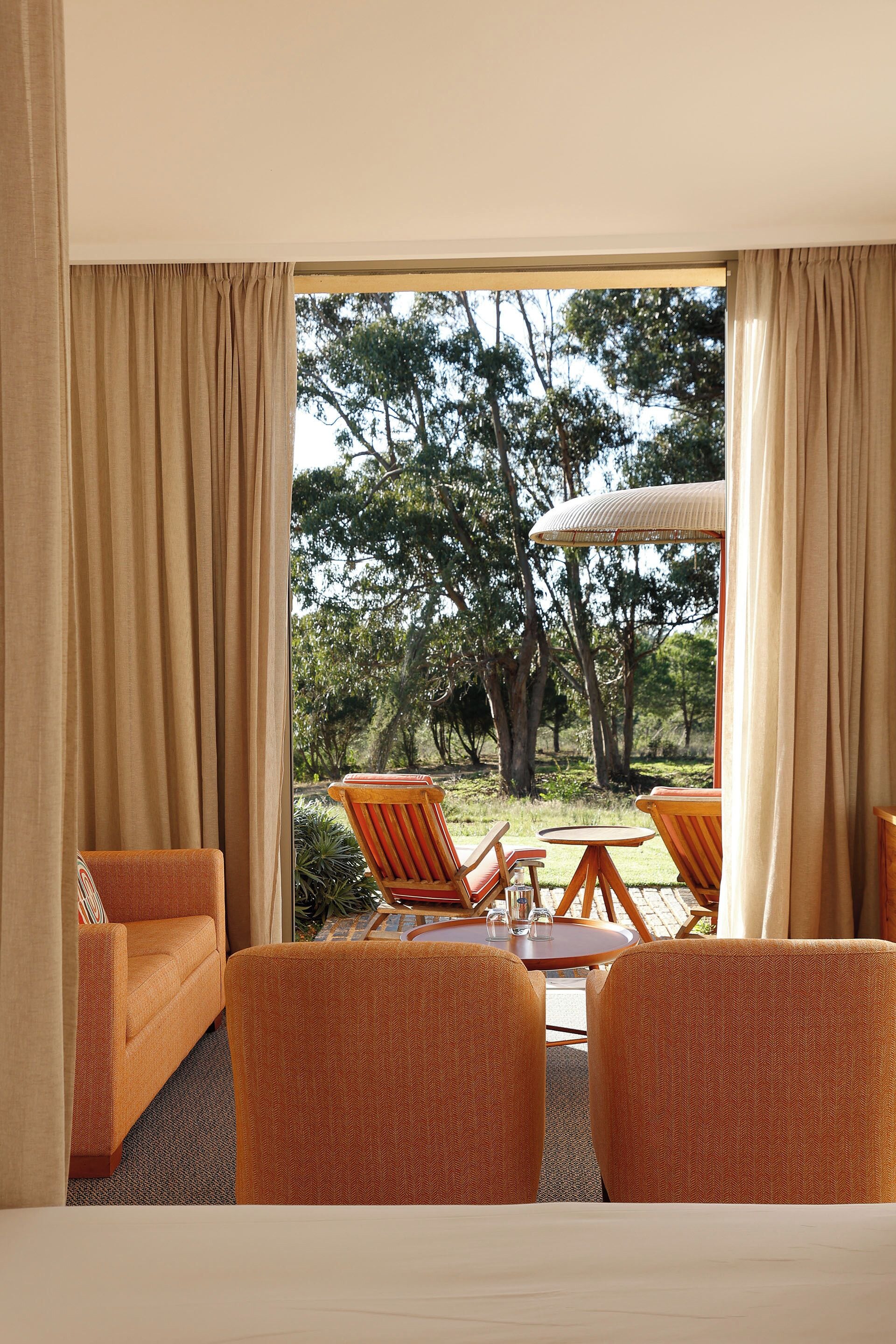 Une chambre d’hôtel avec une grande fenêtre, deux fauteuils orange, une petite table ronde et un lit.
