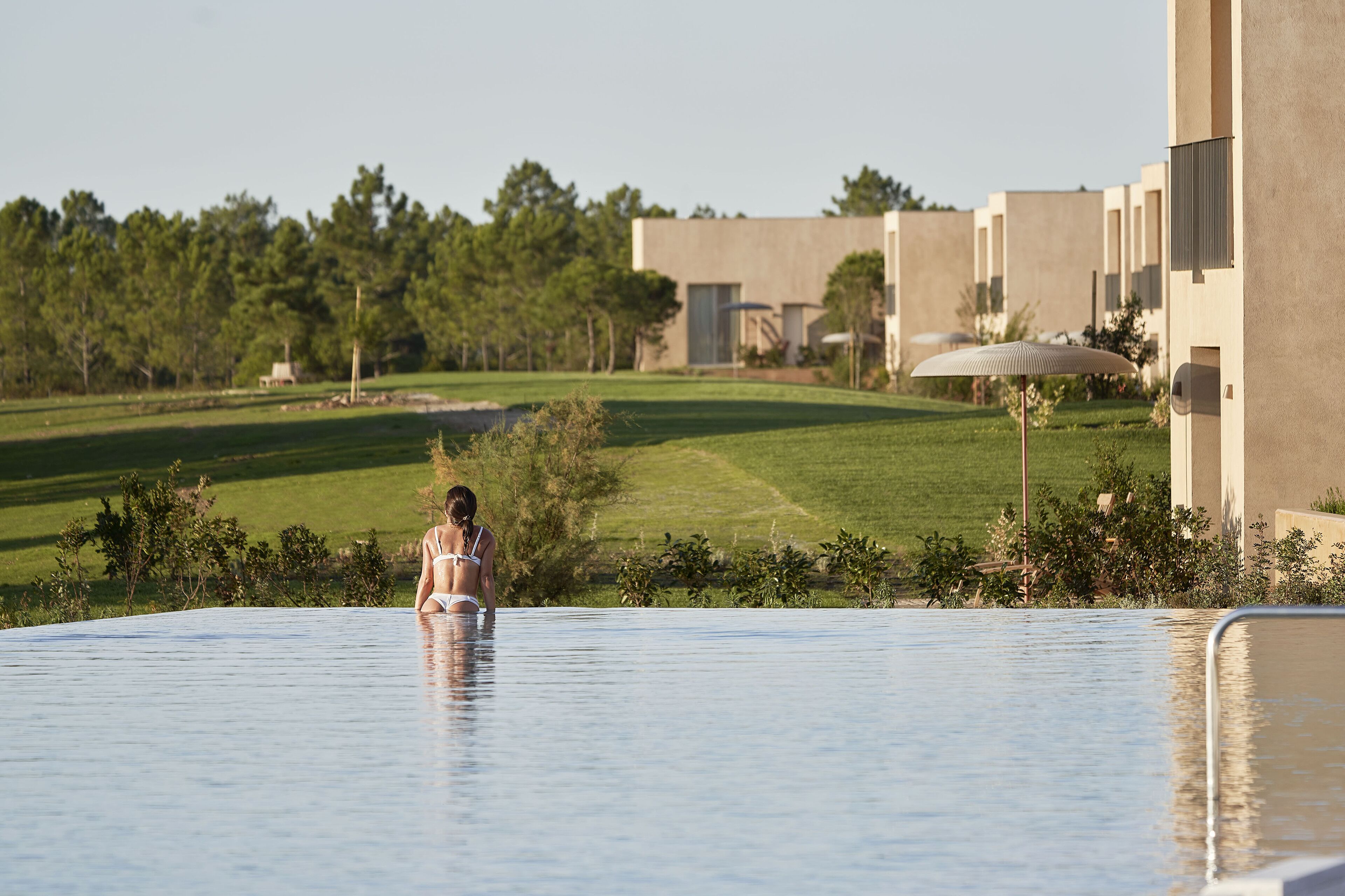 Eine Frau in einem Badeanzug in einem großen, klaren Pool mit Blick auf einen Golfplatz und moderne Gebäude im Hintergrund.