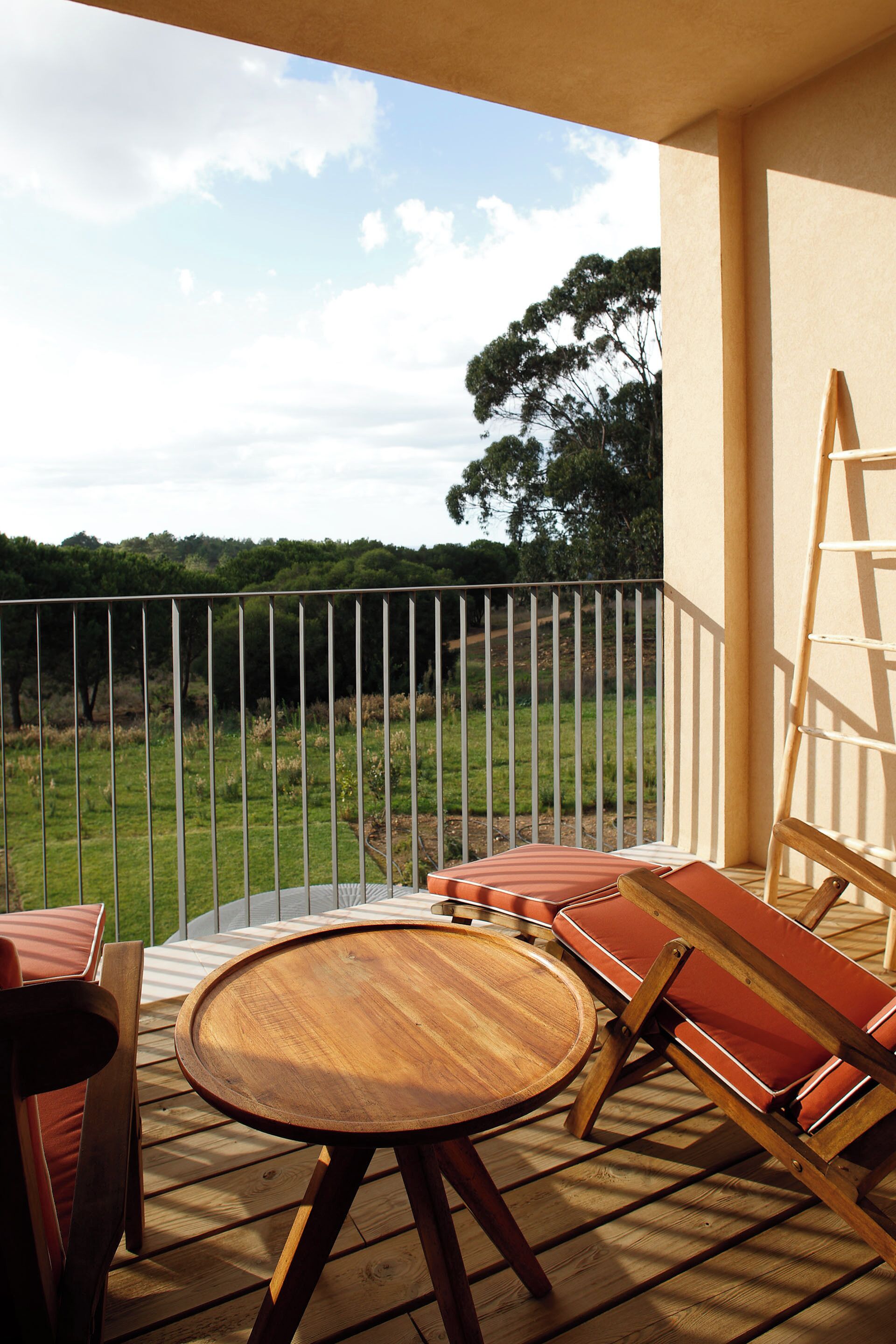 Una terraza de madera con una mesa redonda y sillas acolchadas, con vistas a un paisaje cubierto de hierba y árboles.