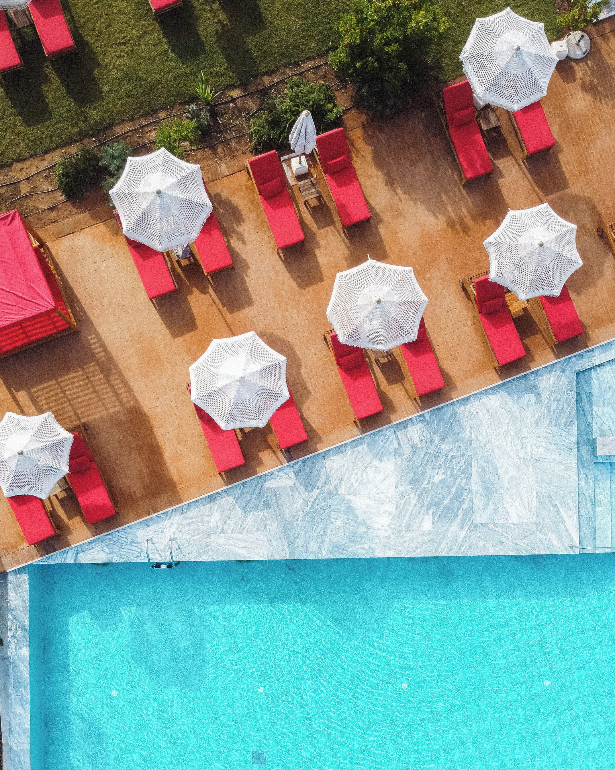 Vista aérea de una zona de piscina con múltiples sillones rojos y sombrillas blancas.