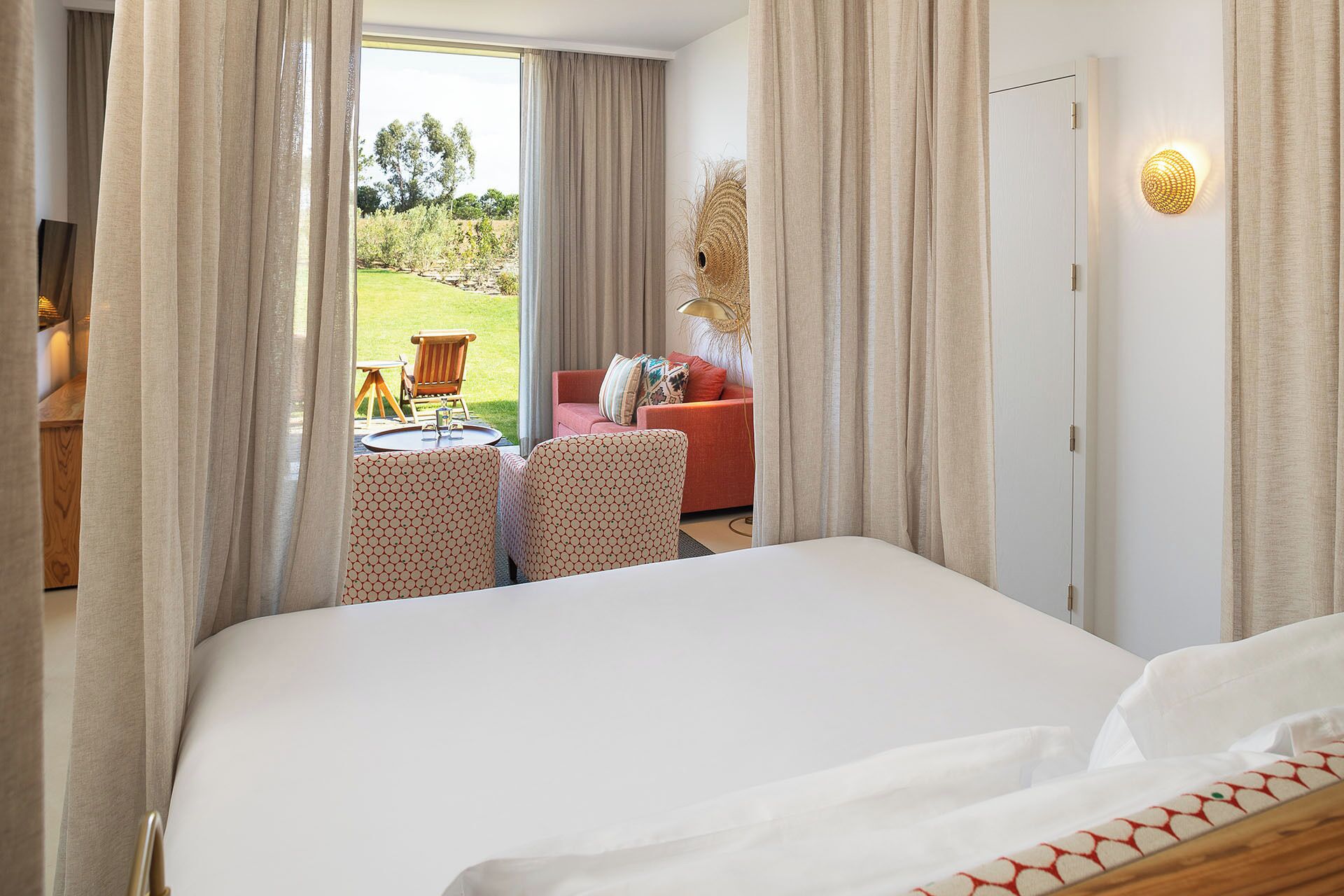 A hotel room with a large bed, a red sofa, two patterned armchairs, and a view of an outdoor garden area through a glass door.