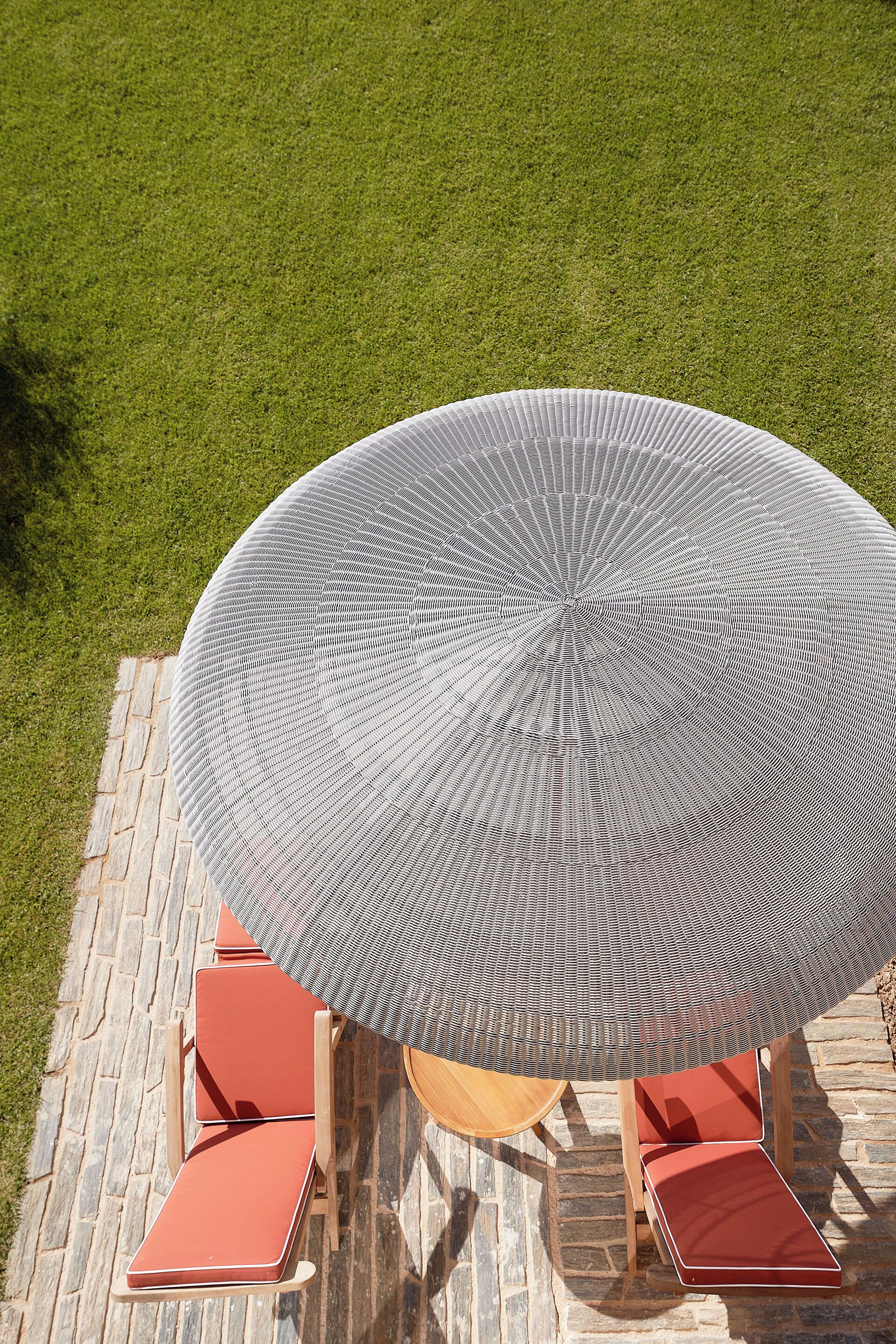 Une table ronde surmontée d’un parasol à motifs, entourée de chaises rouges à coussins, le tout posé sur une terrasse en pierre.