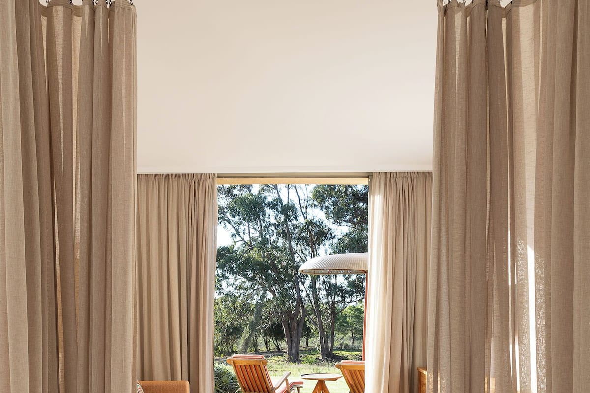 A hotel room with a large bed, beige curtains, and a view of an outdoor seating area with chairs and a table.