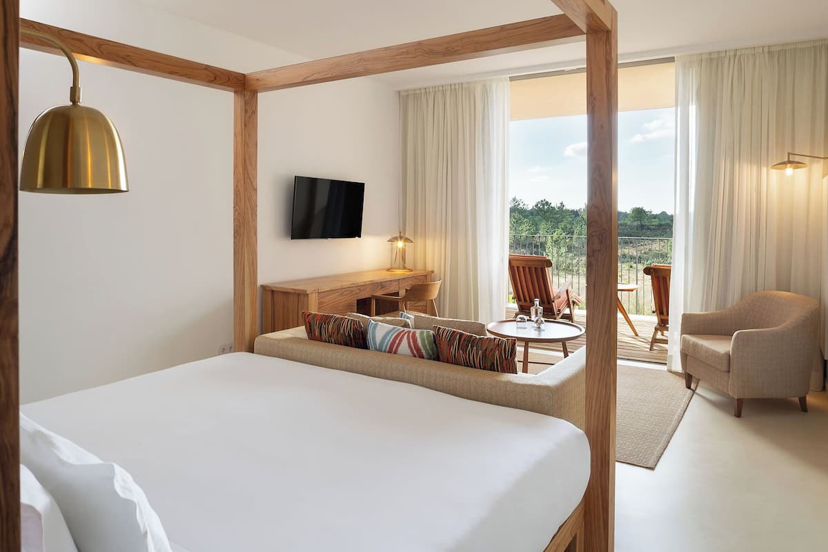 A hotel room with a large bed, a desk with a chair, a small table with a chair, and a balcony with a chair.