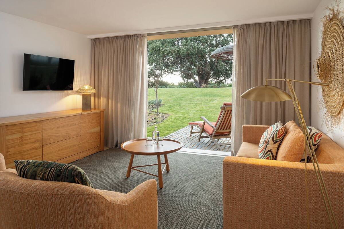 A hotel room with a sofa, a small table, a TV, and a view of the garden through the sliding doors.