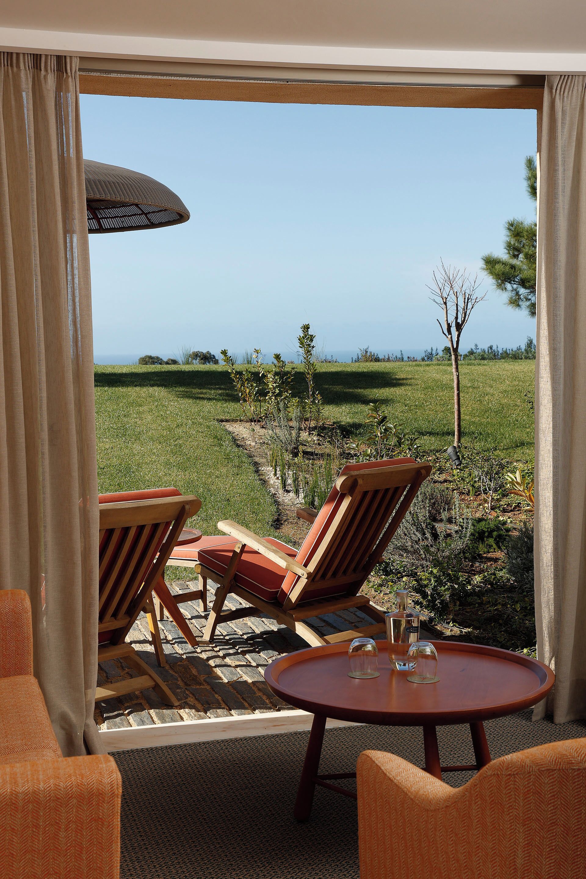 A room with two wooden chairs and a small table, a large umbrella, and a view of a grassy field and trees.