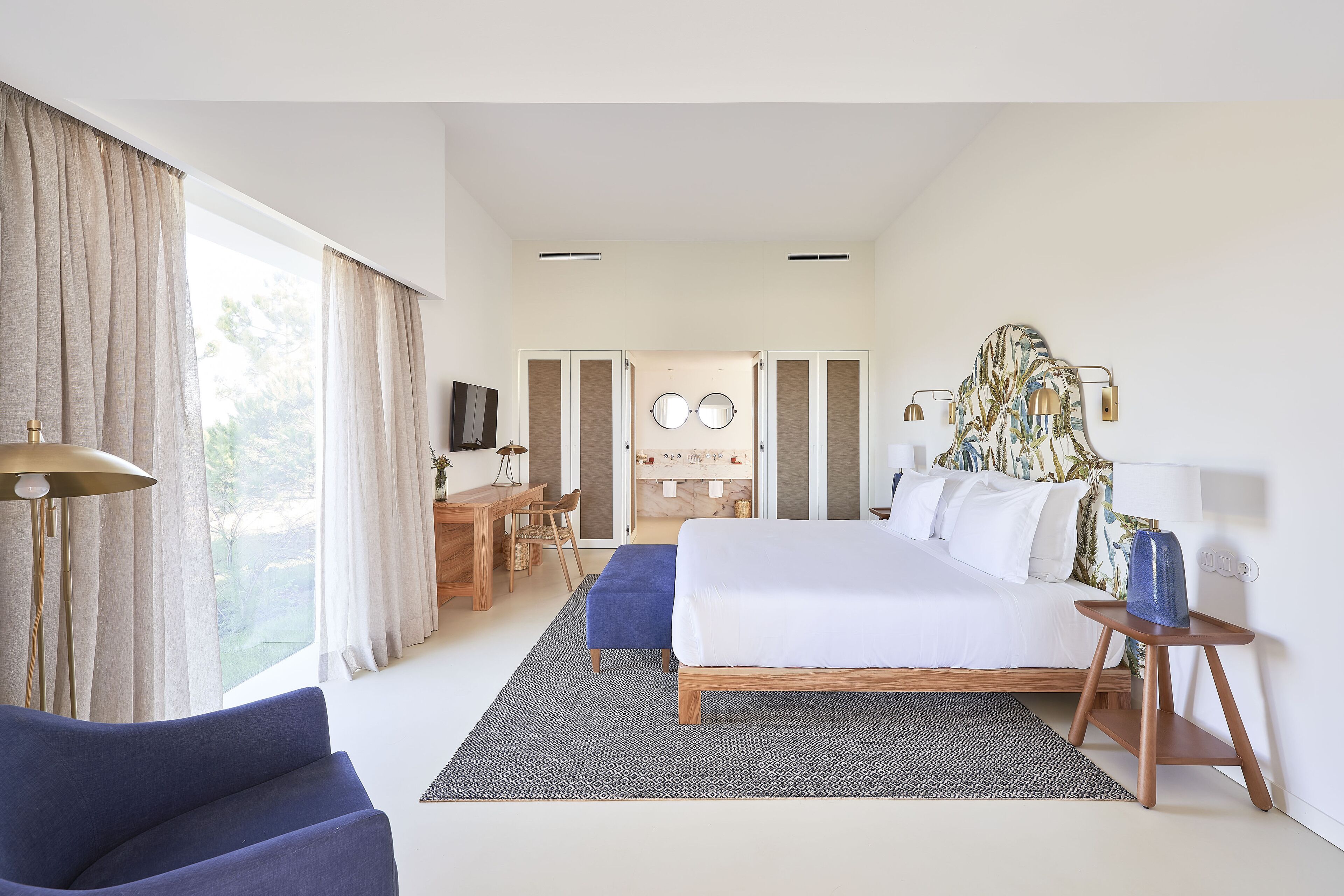 A modern hotel room with a large bed, a desk, and a chair. There is a view of the outdoors through large windows.