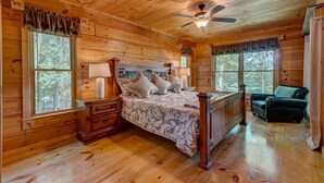 A wooden cabin bedroom with a four-poster bed, two bedside tables, a chair, and large windows. - Poppy's Place - Mountain View | Hot Tub | Fire Pit | Shuffleboard | Ping Pong (Ellijay)