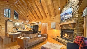 A cozy log cabin interior with a stone fireplace, wooden floors, a leather sofa, and a dining area. - Poppy's Place - Mountain View | Hot Tub | Fire Pit | Shuffleboard | Ping Pong (Ellijay)