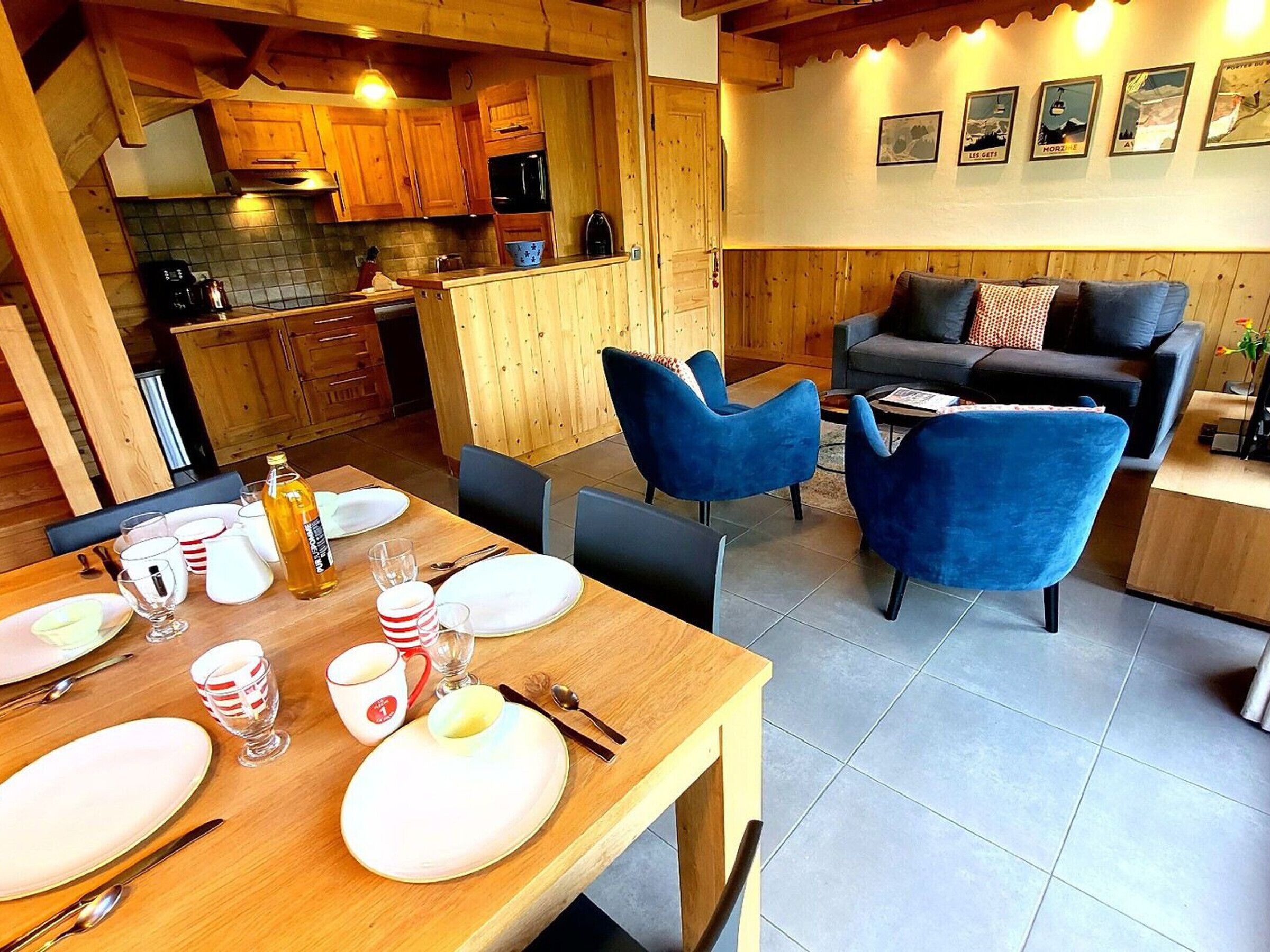 Um quarto acolhedor com uma mesa de jantar de madeira posta para dois, um sofá com cadeiras azuis, e uma cozinha com armários de madeira e painel de azulejos.