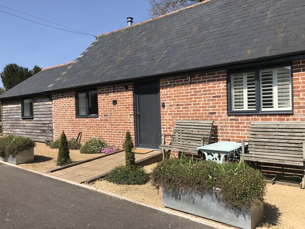 Un bâtiment en briques avec un toit gris, une porte noire et une fenêtre. On y trouve deux bancs en bois et une table à l’extérieur.