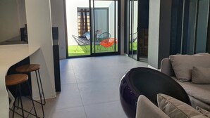 A modern living room with a large sliding glass door leading to a balcony. The room features a sofa, a coffee table, and a kitchen area with stools. - Architect-designed villa with heated pool (Moliets et Maa)