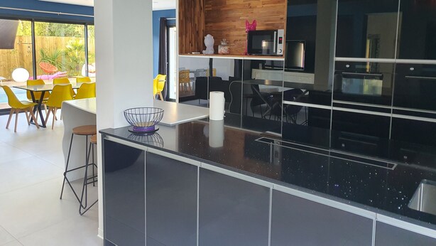 A modern kitchen with black cabinets, a wooden accent wall, and a dining area with yellow chairs. - Architect-designed villa with heated pool (Moliets et Maa)