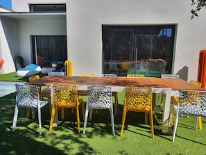 An outdoor dining area with a wooden table and chairs, surrounded by artificial grass and a pool.