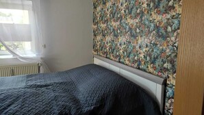 Une chambre à coucher avec un lit, une tête de lit à motifs floraux et une fenêtre ornée de rideaux légers.