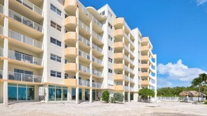 Un hôtel à plusieurs étages doté de balcons, avec un rez-de-chaussée recouvert de sable et un ciel d’un bleu limpide.