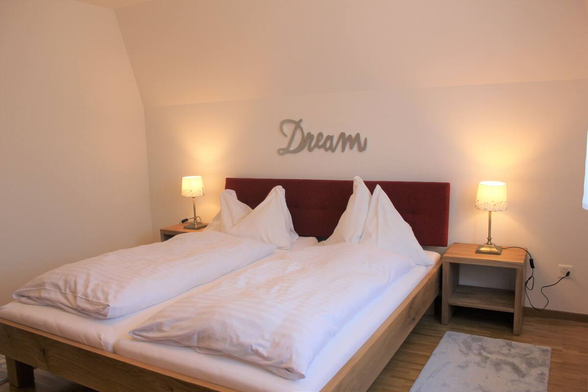 Ein ordentlich bezogenes Doppelbett mit weißen Laken, zwei Nachttische mit Lampen und ein an der Wand befestigtes Schild mit der Aufschrift 'Dream'.