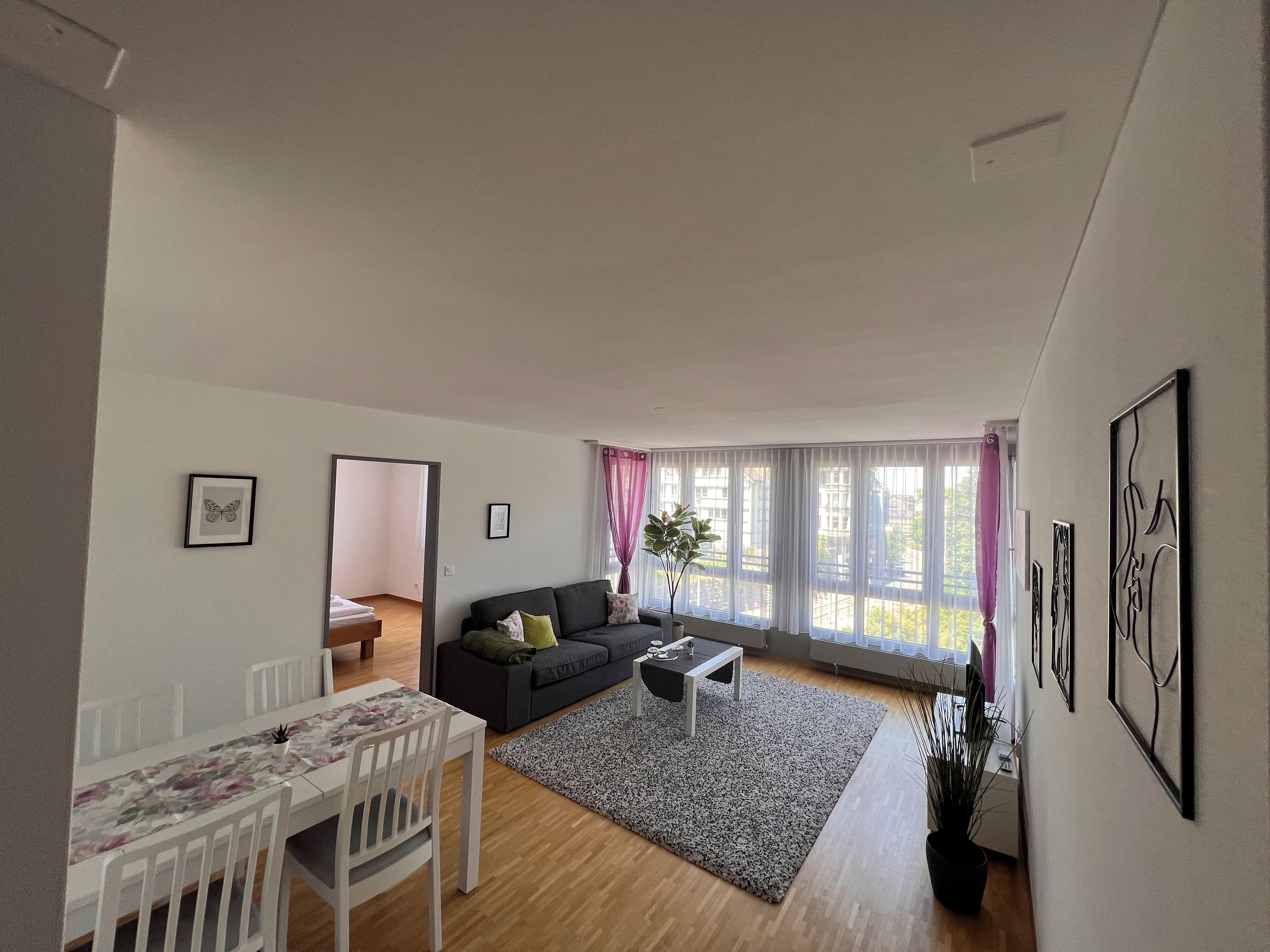 A modern living room with a sofa, coffee table, and dining table. There are large windows with curtains, a mirror, and framed artwork on the walls.