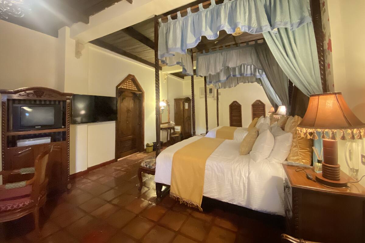 Ein traditionell eingerichtetes Schlafzimmer mit einem Himmelbett, einem Fernseher, einem Schreibtisch mit Lampe und einem Holzstuhl.