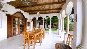 Une terrasse couverte avec une table et des chaises en bois, des piliers blancs et un ventilateur de plafond.