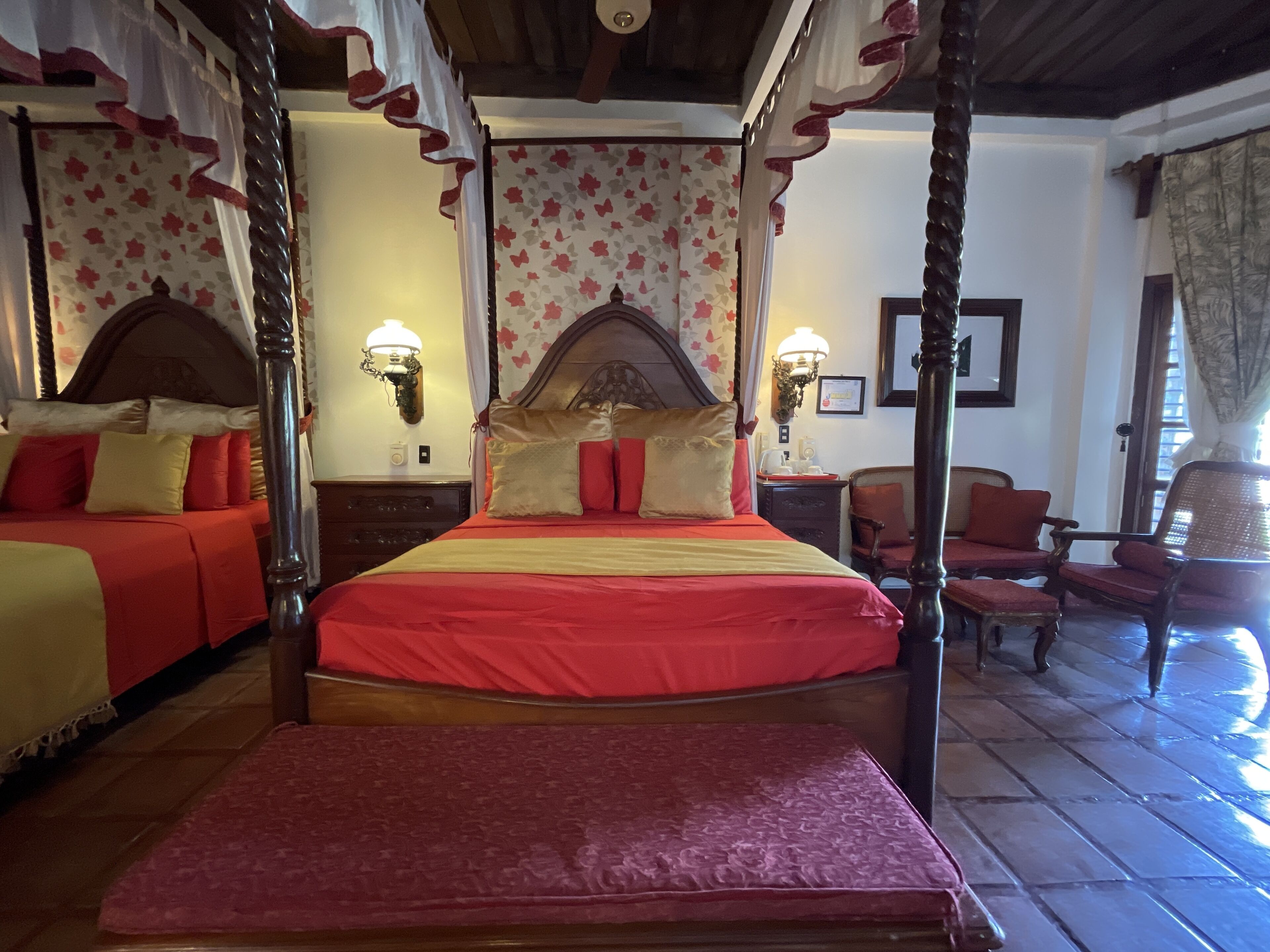 A four-poster bed with a canopy, a wooden headboard, and a red bedspread. There are two bedside tables with lamps, a chair, and a bench. The room has floral wallpaper and a tiled floor.