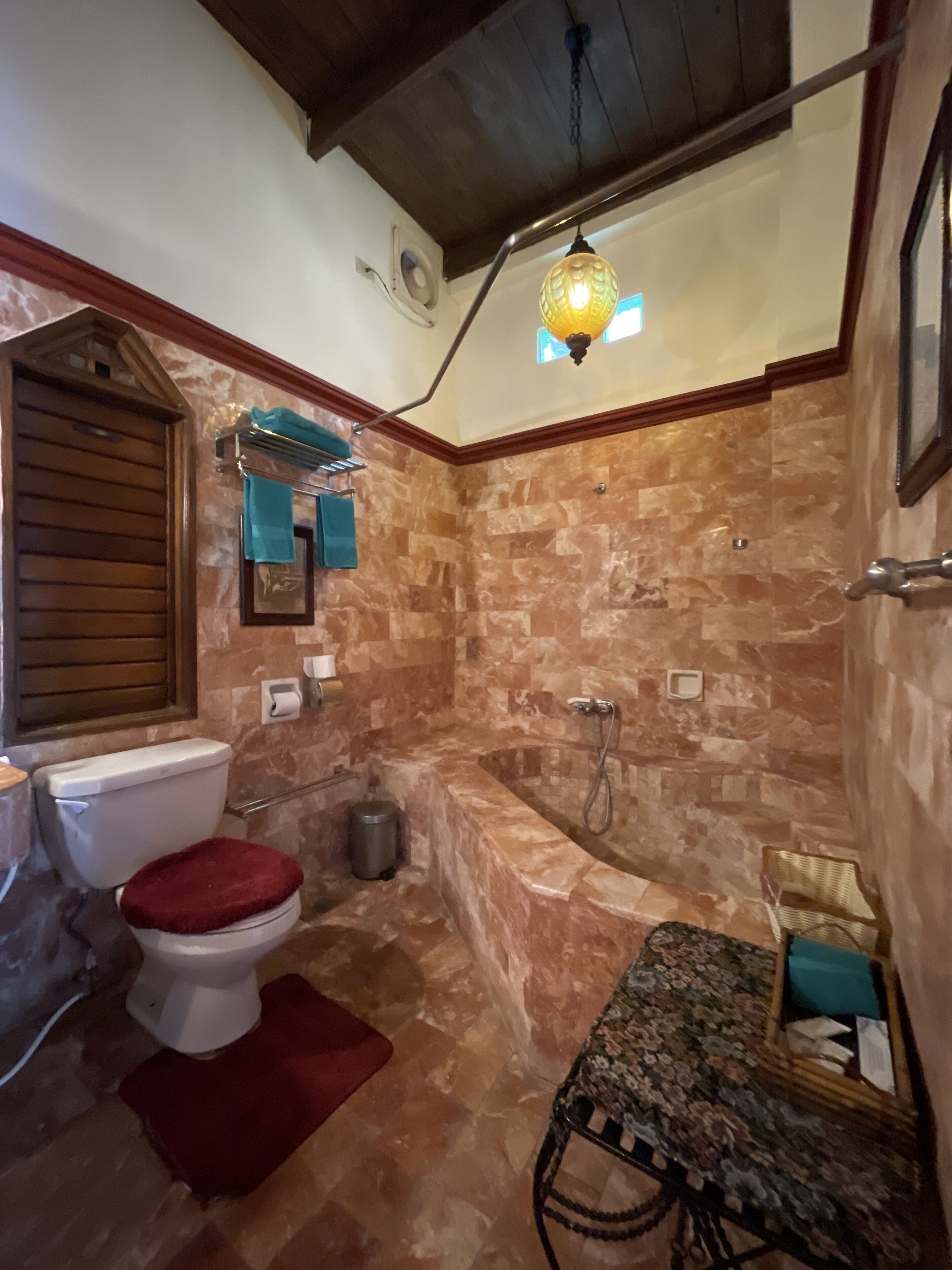 Un baño con pared de mármol, una bañera, un inodoro con el asiento rojo, una lámpara colgante y un pequeño banco con estampado floral.