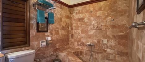 Un baño con pared de mármol, una bañera, un inodoro con el asiento rojo, una lámpara colgante y un pequeño banco con estampado floral.