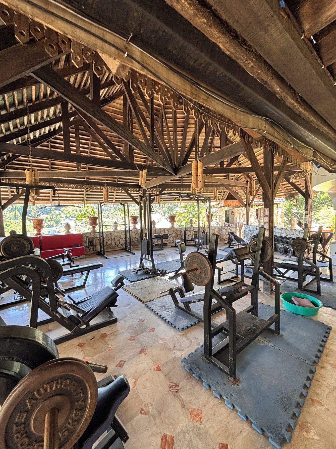 An outdoor gym with various exercise equipment, including weight machines and free weights, under a wooden structure.